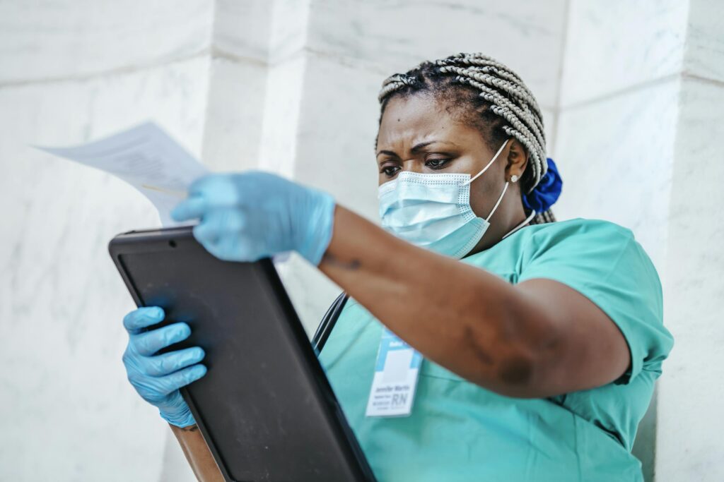 low-angle-of-focused-black-female-nurse-in-mask-and-gloves-standing-in-hospital-and-looking-through-papers-6098056-scaled